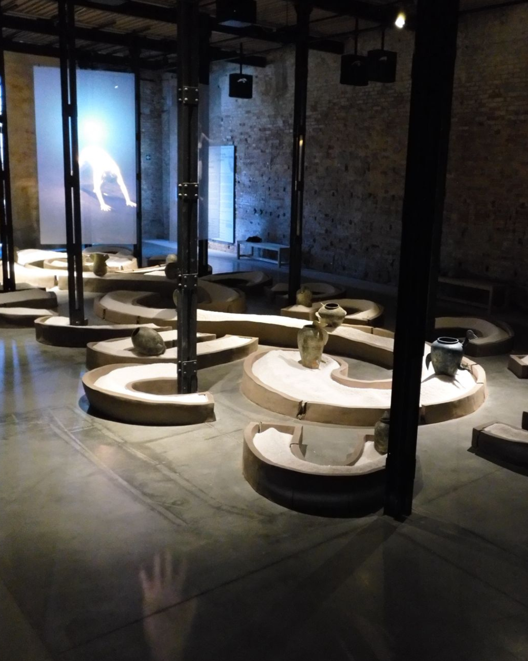 2 of 5, A dark room with a low snaking sculpture around the floor, filled with salt. There are ceramic vases sitting in the sculpture.